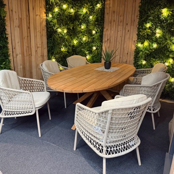 Round wooden table with wicker chairs in an outdoor setting with greenery and lights.