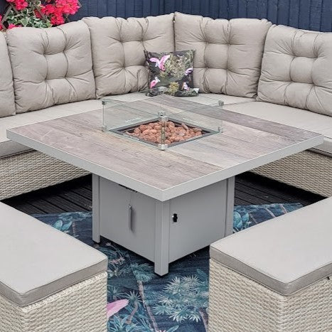 A square gas firepit table with a ceramic tabletop, placed outdoors with a grey sofa and a cushion with a floral design in the background.