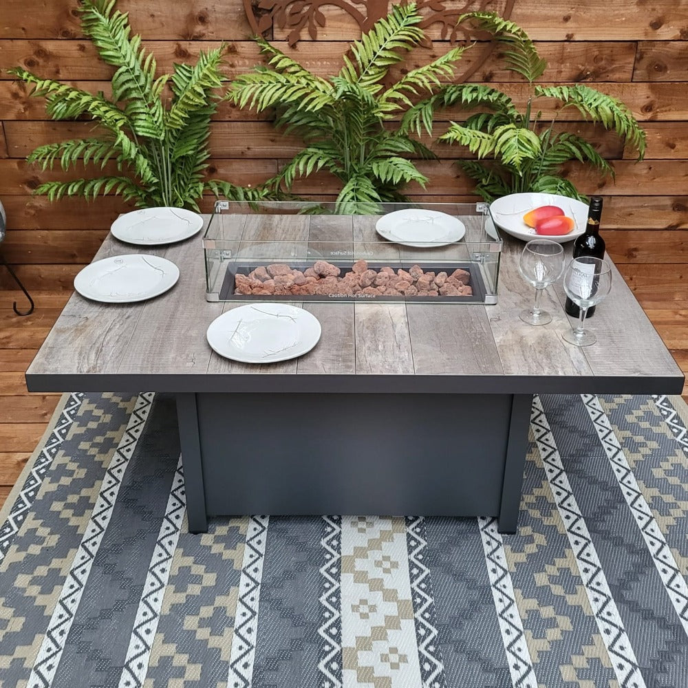 A rectangular gas firepit table with a ceramic tabletop and a metal frame, surrounded by plates and glasses on a patterned rug, indicating it is designed for outdoor relaxation and heating.