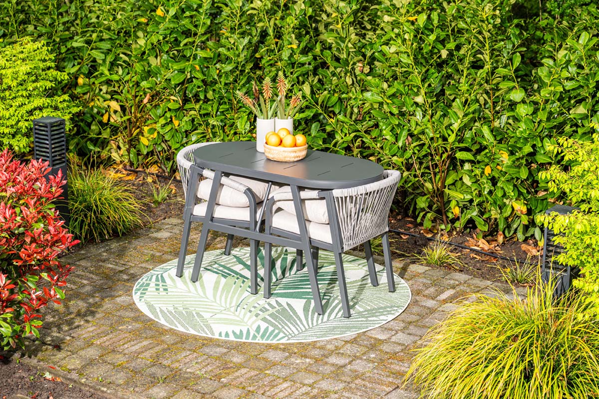 Outdoor patio set with table and chairs on a circular rug in a garden setting.