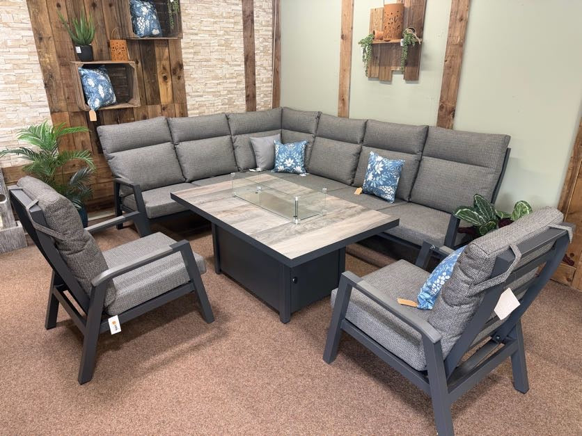 Outdoor furniture set with gray sectional sofa, chairs, and fire pit table against a wooden wall.