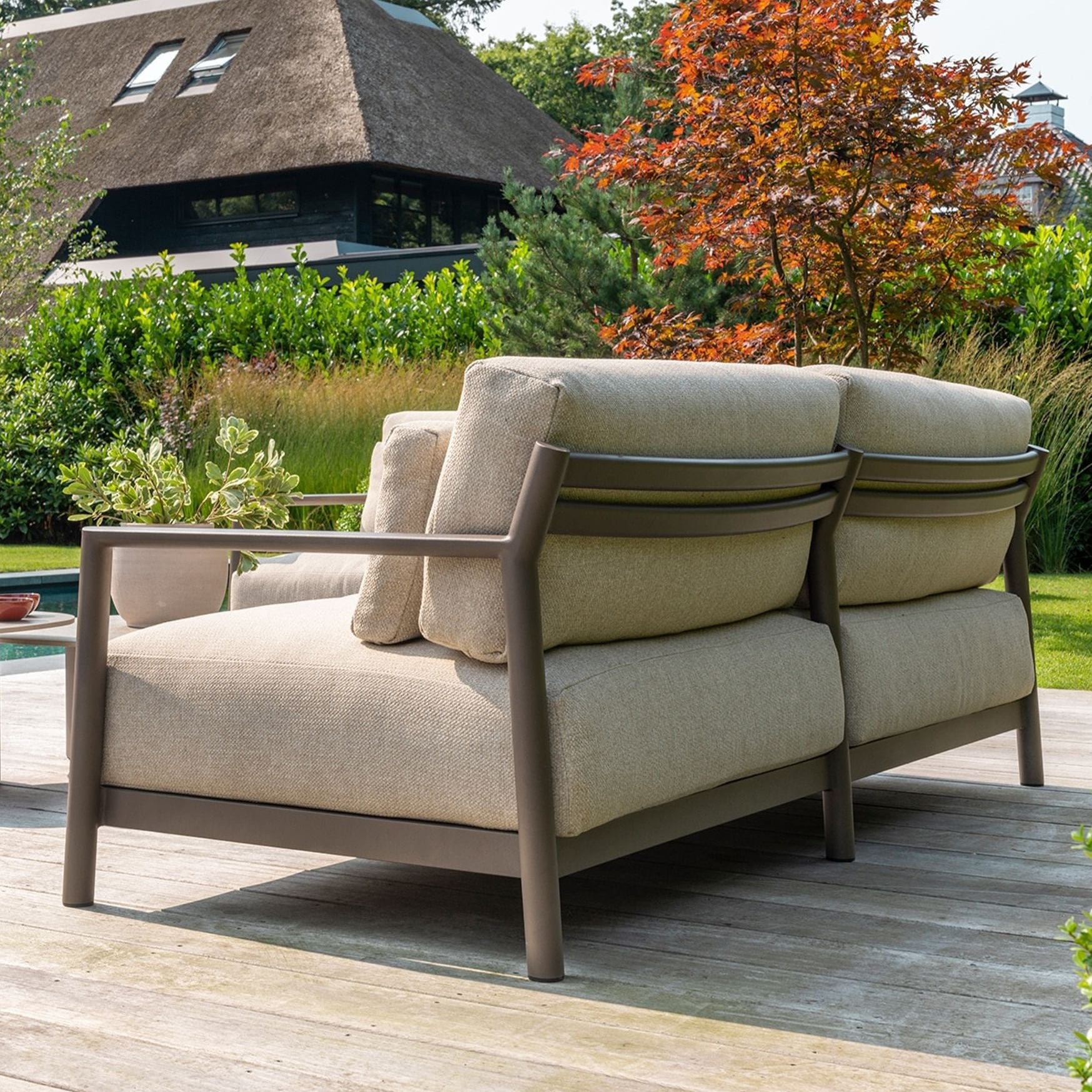 Beige outdoor sofa with cushions on a wooden deck, surrounded by greenery and a pool.
