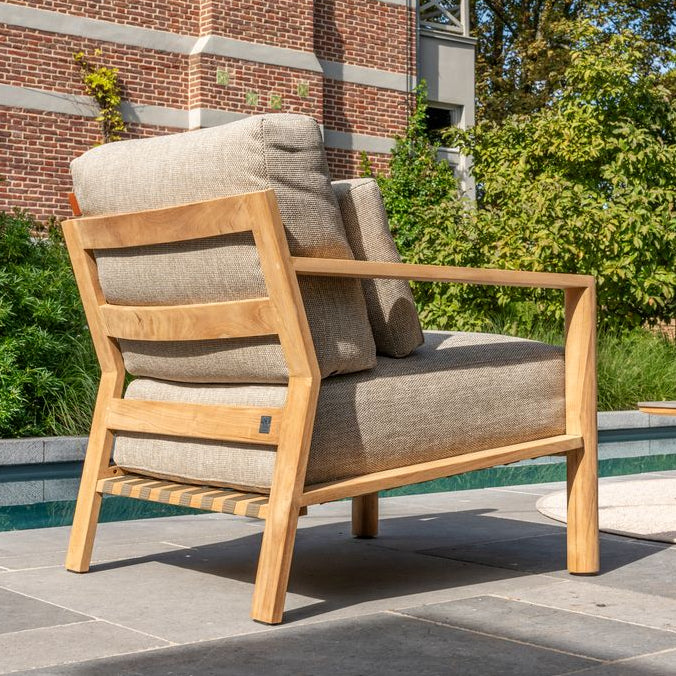 Wooden armchair with gray cushions on a patio area with a brick building and trees in the background.