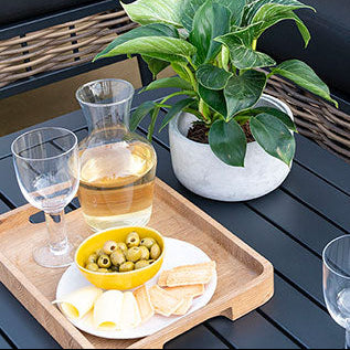 Outdoor patio setting with dark cushions, yellow pillows, a plant, and a tray with snacks and drinks.