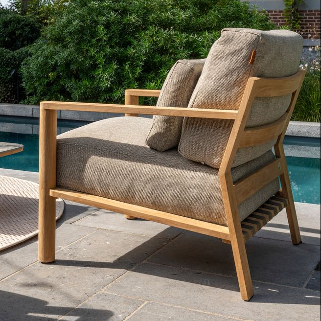 Wooden armchair with beige cushions on a patio by a poolside, surrounded by greenery.